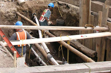 A photo of workers in high-visibility vests and blue hard hats working in a dirt pit with contemporary and wood pipes laid near one-another underground.