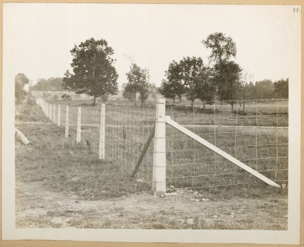 A black and white photo of a country setting. A corner of a fence is in the foreground with one side of the fence extending into the far distance on the left. The fence is made from timber posts and rectangular wire fencing.