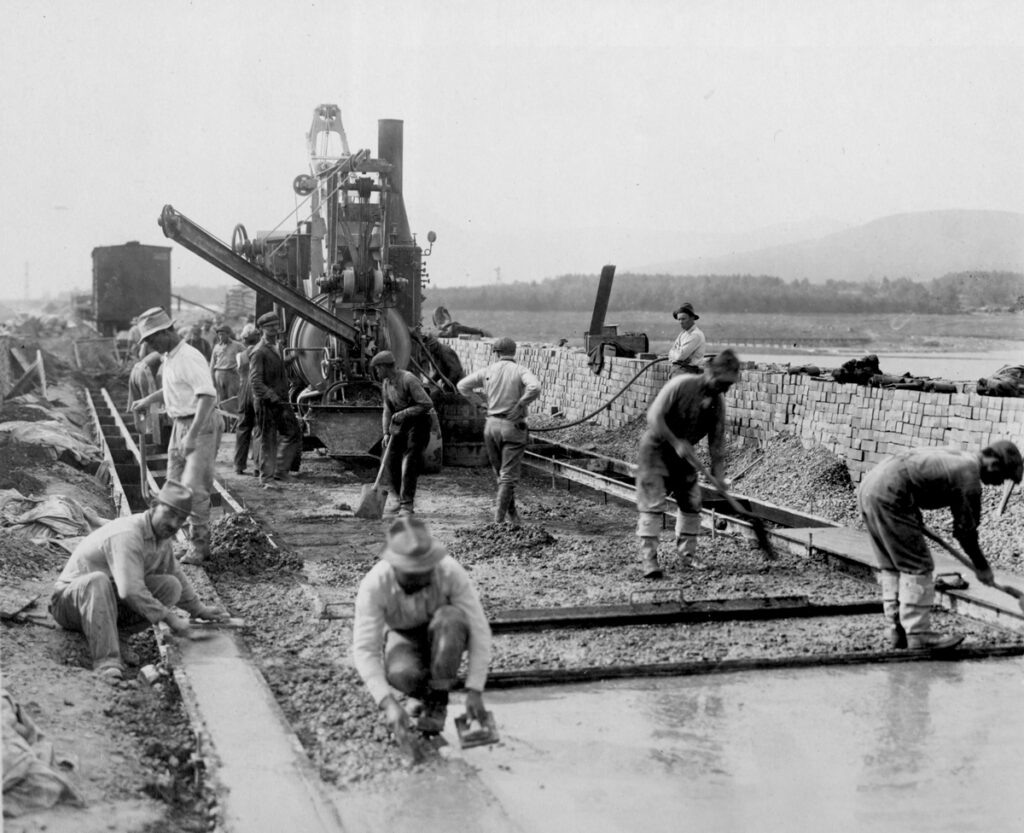 A black and white photo of laborers smoothing down concrete slabs. There is a machine in the background with workers working around and in front of it. A waist height brick wall separates the work from the background landscape which includes a body of water and a distant hazy mountain.