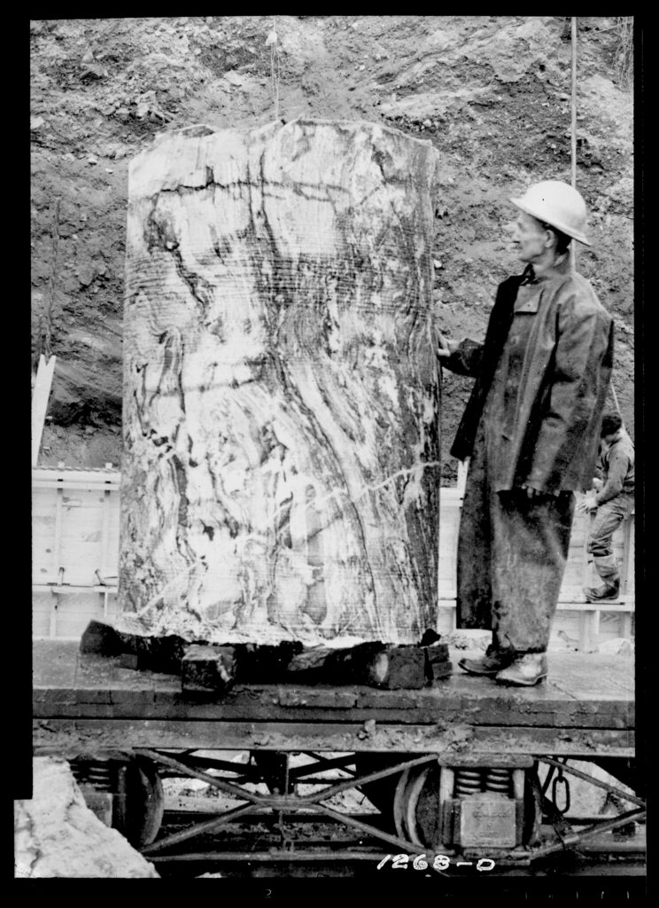 A black and white photo of a man standing next to a column of rock that is taller than he is. It is about 4 ft wide.