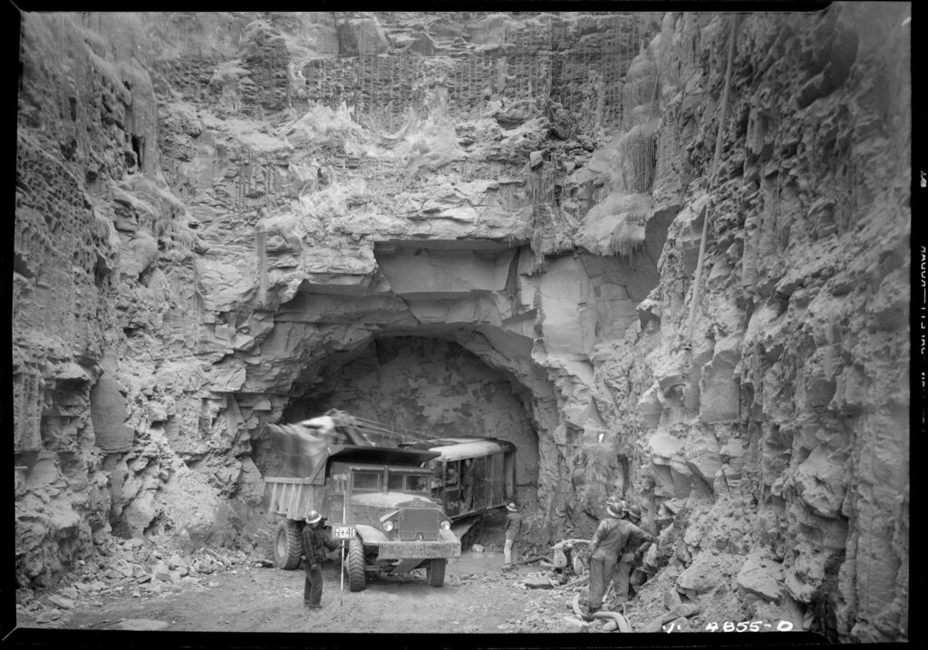A black and white photo from the bottom of an excavated site. Rock has been blasted and carved away to make a large passage leading into the entrance of a tunnel. There is a couple of trucks at the entrance to the tunnel. 