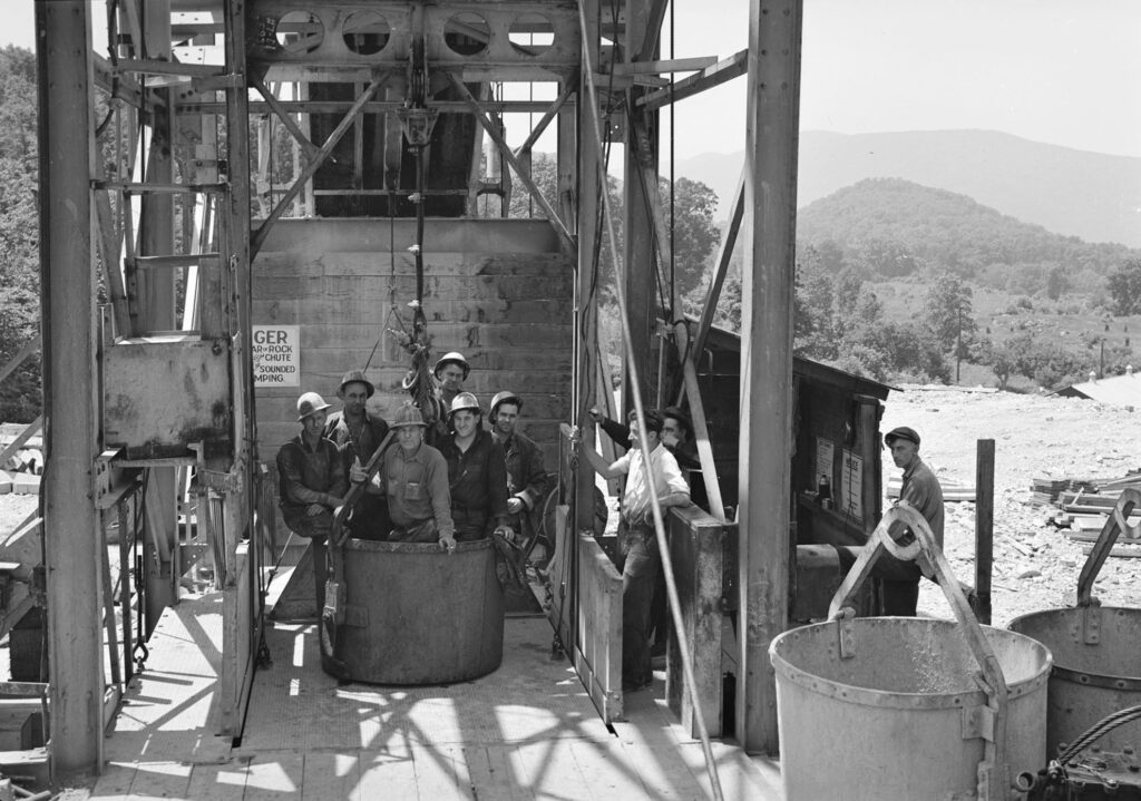A black and white photo of 6 men in hard hats sitting in a metal bucket that is staged on a crane platform. In the background is a hilly landscape.