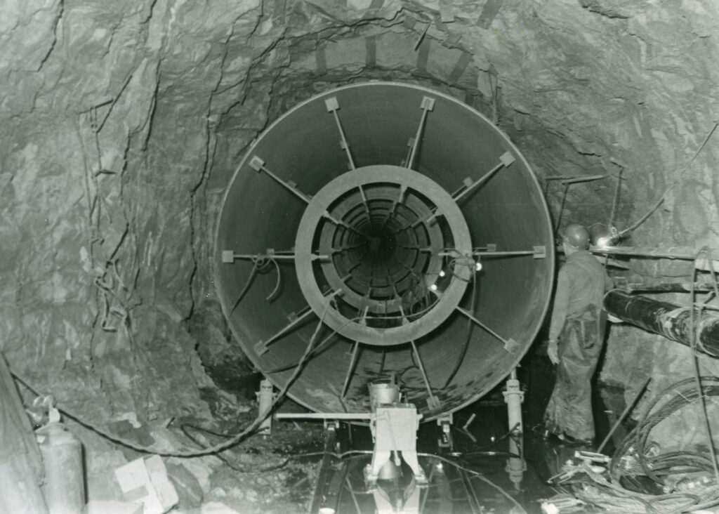 A black and white rock tunnel with a metal tube that fits into the tunnel with just a couple of feet to spare. A man in a hard hat is looking at the tunnel, which is twice his size.