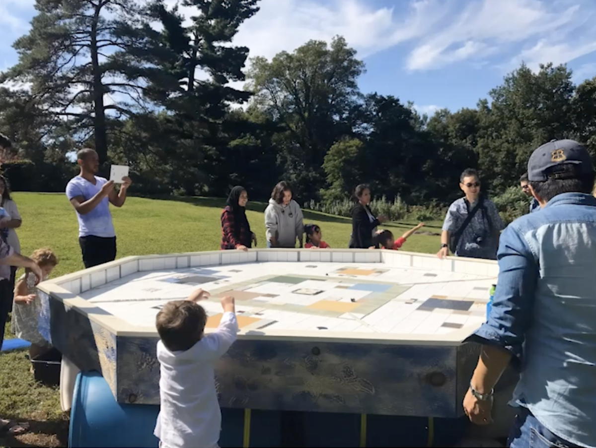 A photo of a sculpture that looks like a white octagonal table with patterns laid onto the surface. It is placed in a field with people and children gathered around it.