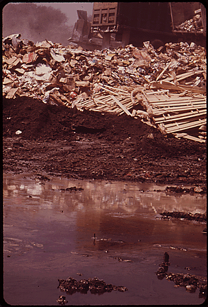 A color photo of a muddy puddle in front of piles of trash.