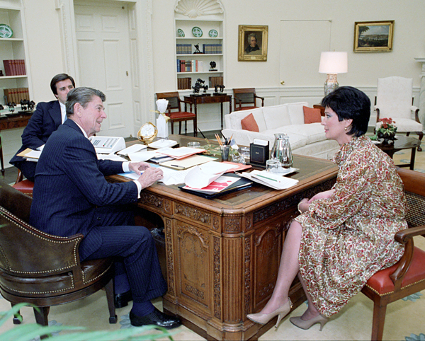 A color photograph of Ronald Reagan, Anne Gorsuch, and another man sitting around the President's table in the Oval Office. Anne Gorsuch wears clothes, hair, and makeup in the style of the 80s, with a paisley pattern dress, shiny stockings, and tan colored high heels.