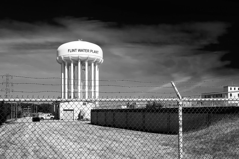 A black and white landscape photo of a white, with a tank sitting on stilts. The words "Flint Water Plant" are written in black on the tank. A chainlink fence lined at the top with barbed wire stands in front of the scene.