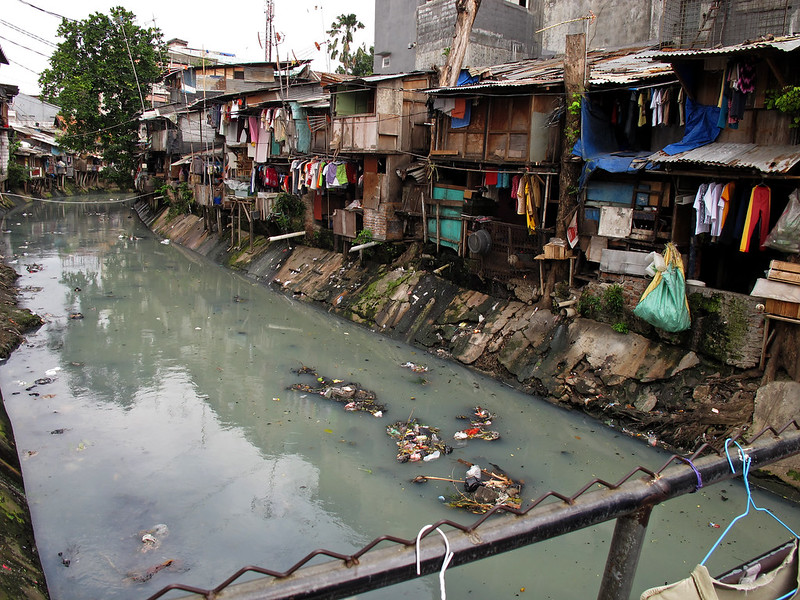 A photo taken from a bridge overlooking a canal, half-filled with dirty and polluted water. Around the banks are makeshift shacks, constructed from found materials