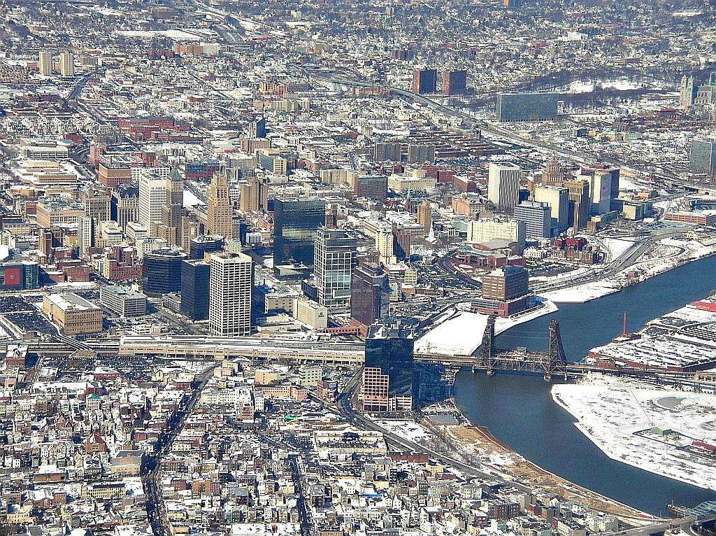 A view from an airplane of an urban city in winter. There is snow on the ground in areas not covered by buildings. The bend of a river cuts through the bottom right of the photo with a rusty bridge connecting the sides of the river.