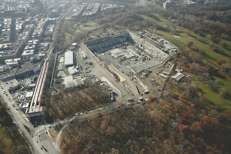 An aerial view of a construction zone, with a big grey concrete hole in the earth surrounded by roads, golf course land, and residential zones.