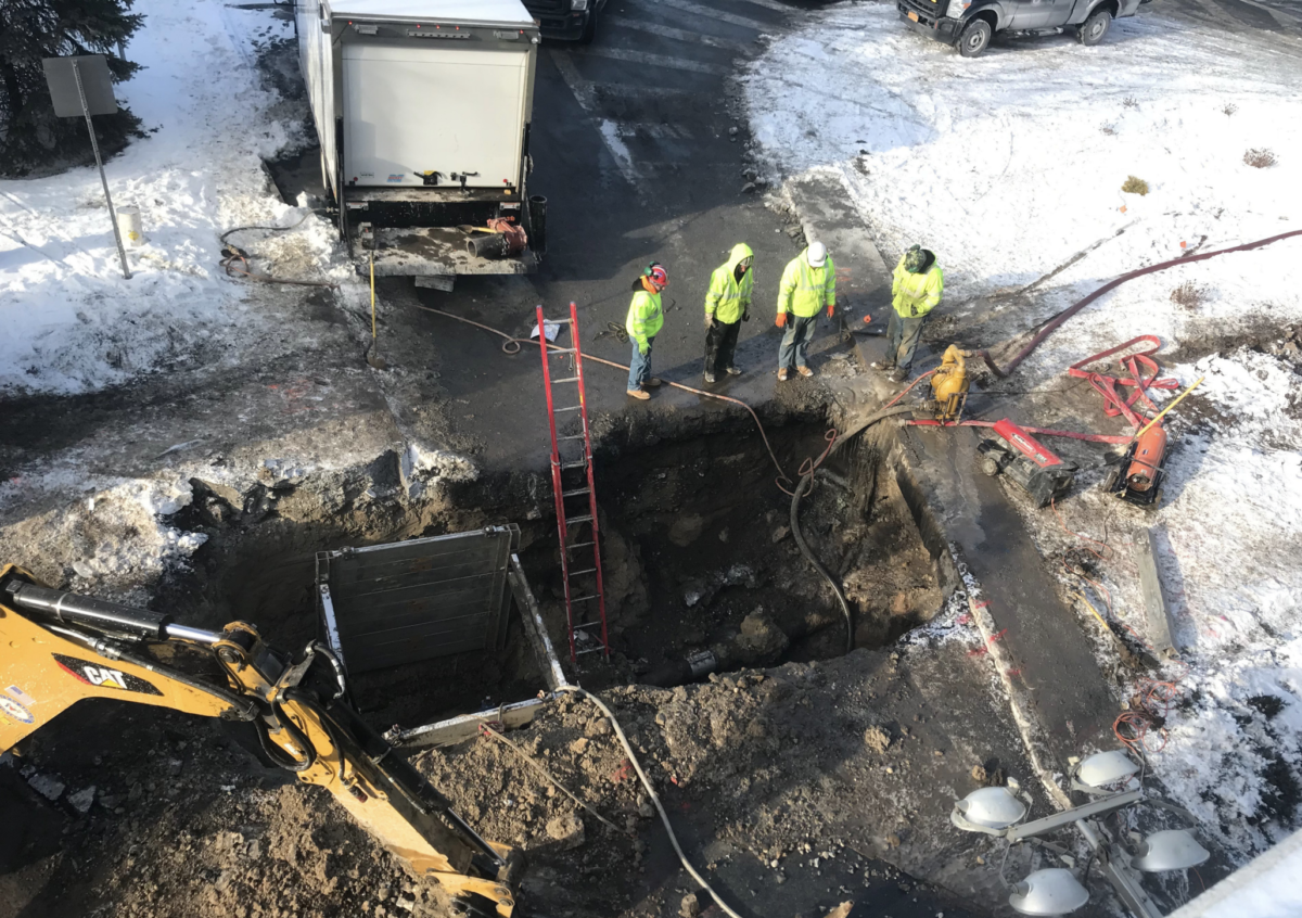 An overhead photo of a large rectangular hole in the ground, about 6ft by 20ft in size. There is snow on the ground and the ground is dirt grey. There's five men in high visibility work wear and hard hats standing around the hole looking around. Tractors and trucks surround the scene.